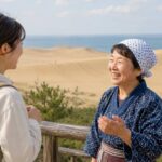 。鳥取砂丘を背景に、旅人が地元の人とやさしく会話しているブログ用アイキャッチ画像